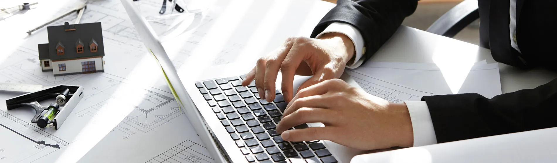 Una persona escribiendo en una computadora personal con unos planos al lado